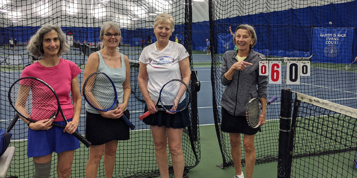 Group of adults posing with tennis rackets in an indoor tennis court.